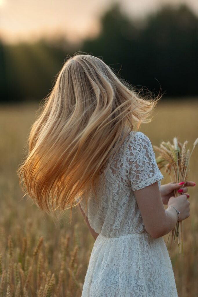 pexels photo 10329696 A woman with flowing blonde hair in a white lace dress stands in a sunlit wheat field holding wheat stalks.